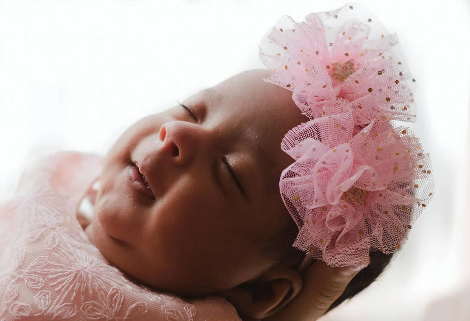 Understanding Baby Sleep Cycles: A close-up of a smiling baby with closed eyes, wearing a pink lace outfit and a pink hair accessory adorned with soft tulle and subtle sparkles, against a light background.