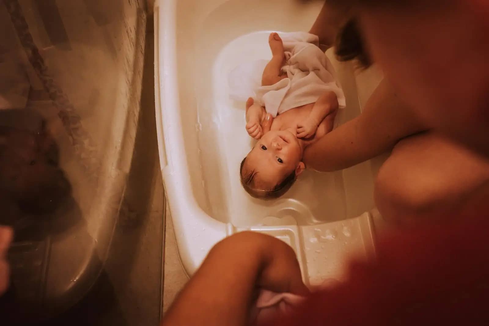 Newborn Bathing Guide: A baby wrapped in a towel is lying in a bathtub partially filled with water, with one arm raised above its head, while an adult's leg is visible beside the baby.