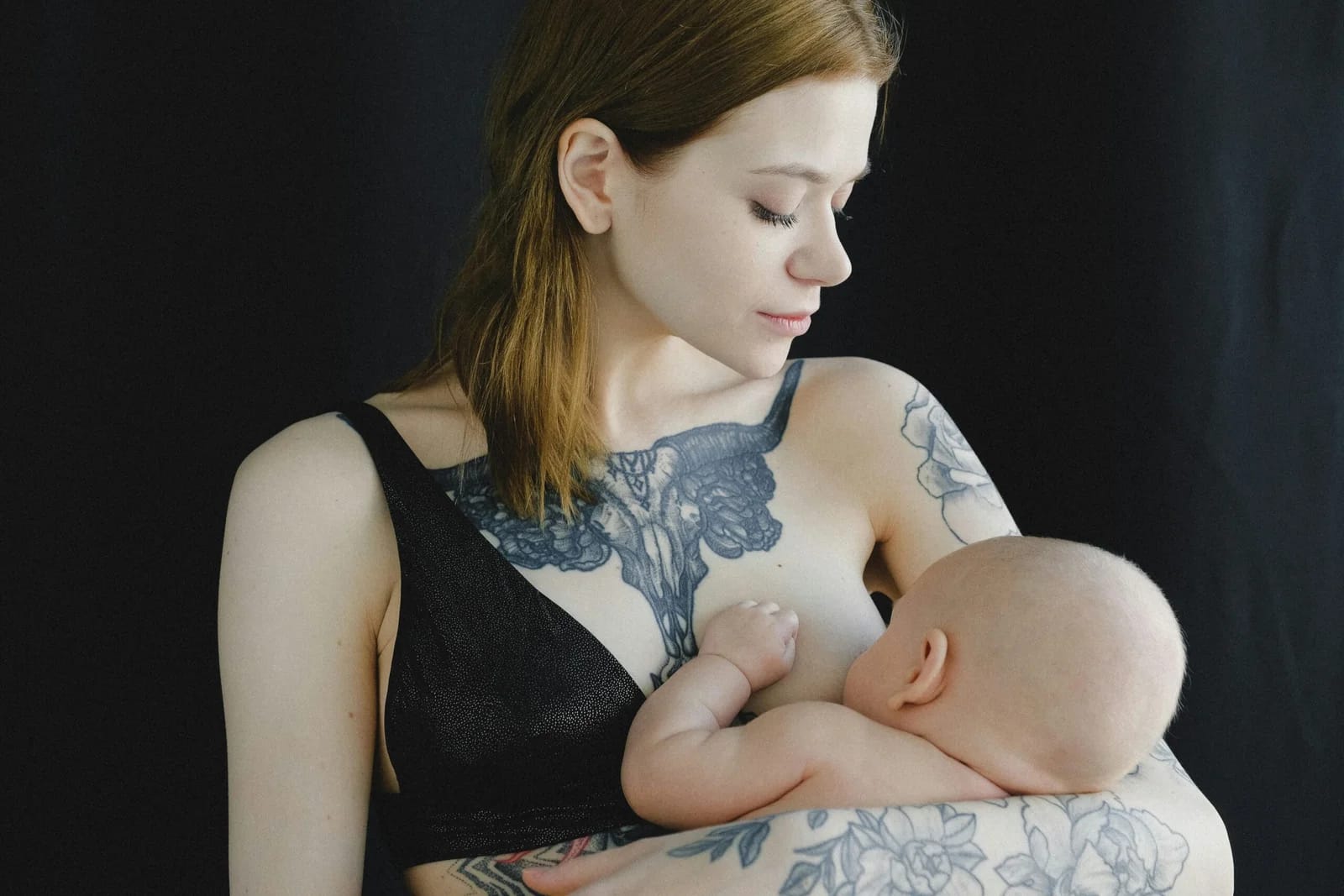 A woman with shoulder-length brown hair and intricate tattoos on her chest and arms is breastfeeding a baby against a dark background.