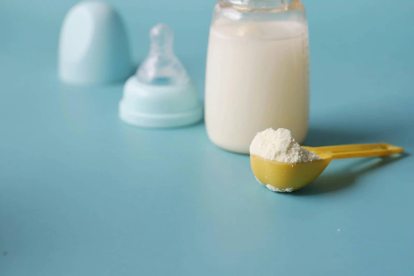 Formula Feeding: A yellow measuring scoop filled with white powdered formula next to a transparent bottle of milk on a light blue surface with a baby bottle cap in the background.