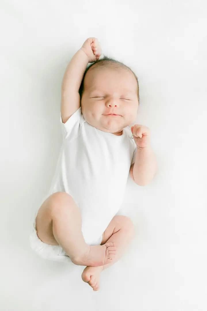 A smiling baby in a plain white onesie is lying on a soft white surface, with one arm raised confidently and the other hand clenched in a fist.