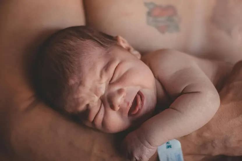 How to Soothe a Crying Baby: A newborn baby with a facial expression indicating discomfort or crying, resting on the arm of an adult who has a tattoo visibly displayed on their skin, capturing a moment of tender vulnerability and connection.