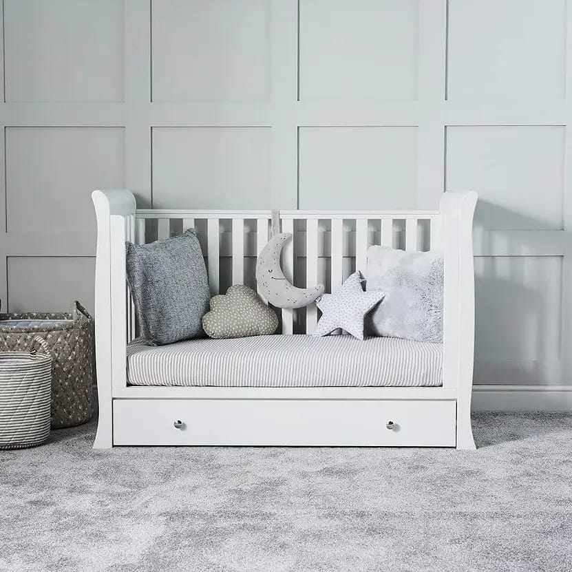 A white wooden daybed with a striped mattress, adorned with various soft pillows including a grey one, a star, a cloud, and a moon, set against a light grey wall with a paneled design.