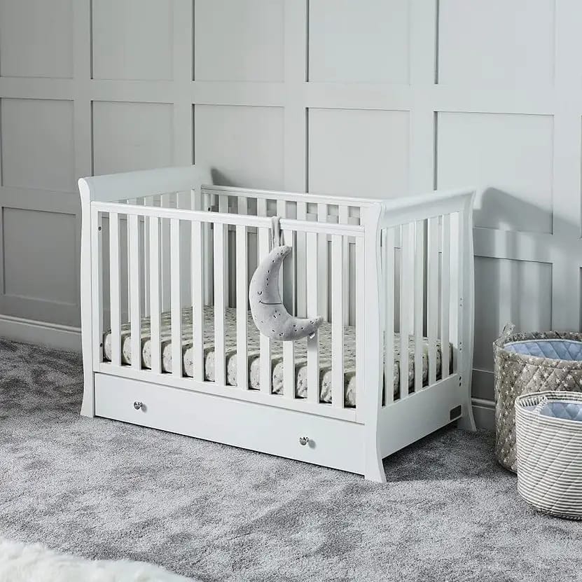 A white wooden crib with slatted sides, featuring a grey patterned mattress, a hanging grey moon decoration, and a storage drawer underneath, set against a light grey paneled wall in a softly carpeted nursery with woven baskets nearby.
